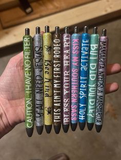 a hand holding five pens with writing on them in different colors and designs, all lined up together