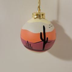 a painted ornament hanging from a gold chain on a white wall with a cactus in the background