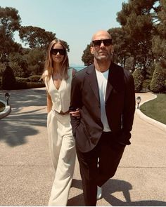 a man and woman are walking down the street in front of some trees with sunglasses on