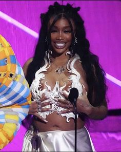 a woman standing in front of a purple background holding a microphone and wearing a white outfit