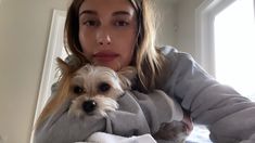 a woman holding a small dog wrapped in a blanket on top of a bed next to a window