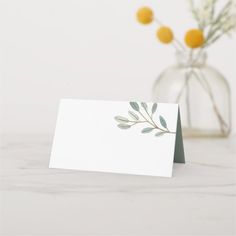 a white card with green leaves on it next to a vase filled with yellow flowers