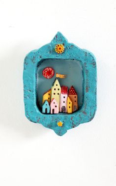 a blue frame with small houses in it on a white wall behind a red and yellow clock