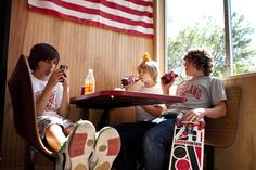 three boys sitting at a table with drinks and skateboards