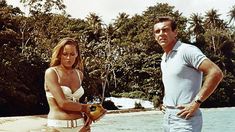 a man standing next to a woman on top of a boat in the water with palm trees behind them