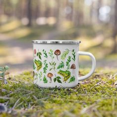 a white coffee cup sitting on top of a green grass covered forest floor in front of trees