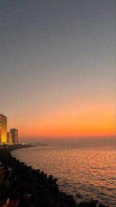 people are sitting on the shore watching the sun rise over the water and buildings in the distance