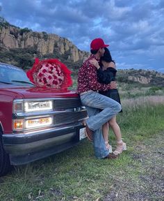 two people sitting on the hood of a car with a red flower in the trunk