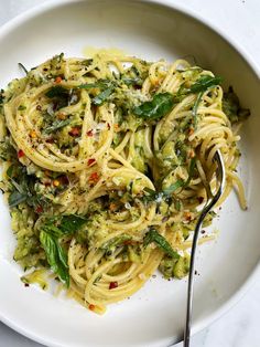 a white bowl filled with pasta and greens on top of a marble table next to a fork