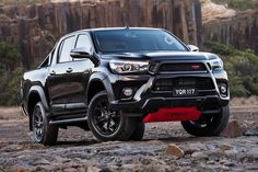 the front end of a black toyota truck parked on rocks near some trees and mountains