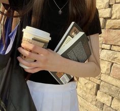 a woman holding coffee cups and books while standing next to a brick wall with her hands in her pockets