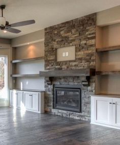 an empty living room with stone fireplace and built - in shelving units, windows, and ceiling fan