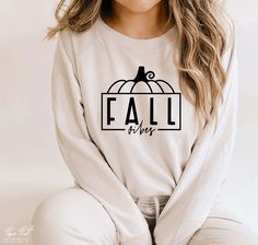 a woman sitting on the ground wearing a white shirt with black lettering that says fall is here