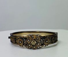 Antique gilt hinged bangle bracelet with floral bouquet and marcasite details, 1940s vintage, dainty details. A geometric like base surrounds the wrist and the front secton has a lovely floral motif with bouquets of sweet flowers. Each of the flowers has a center of a faceted marcasite stone. The clasp and hinge work well, you may need to guide the clasp in gently but it is secure once latched. The color of the piece is an antiqued gold tone or gilt color. There is no makers mark. If you are concerned about fit, we recommend comparing our measurements to a similar item in your collection that you know fits you well.  Specs at a glance Width: 3/4 inches at widest point Interior Diameter: 2.25 Interior Circumference: 6.5 inches We have much more in store and in our showroom than online. Plea Elegant Bangle With Antique Finish, Vintage Hinged Bracelets For Wedding, Victorian Wedding Bracelets In Antique Gold, Victorian Antique Gold Bracelets For Wedding, Vintage Filigree Bracelet For Wedding, Vintage Filigree Bracelets For Wedding, Vintage Metal Bangle For Wedding, Vintage Wedding Bracelets With Antique Finish, Antique Hinged Bracelets For Wedding