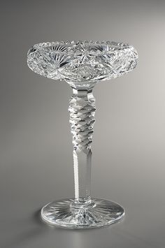 a crystal pedestal with an ornate design on the top and base is shown in front of a gray background