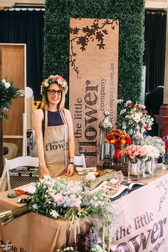 a woman standing in front of a table with flowers on it and a sign that says the little flower shop