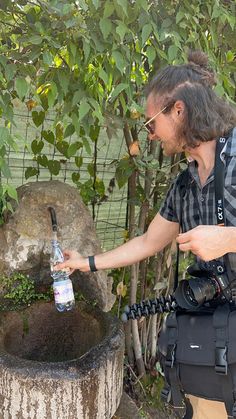 a man holding a camera and looking at an animal in a water fountain with trees behind him