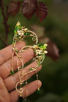 a hand holding a piece of jewelry in it's left hand, with leaves and flowers on the other side