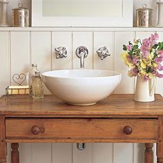 a bathroom sink sitting on top of a wooden table next to a mirror and vase with flowers