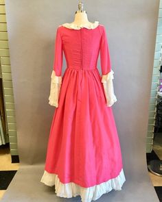 "Lovely reproduction colonial costume in deep, almost hot pink. The low square cut bodice is trimmed in white lace net. The bodice has an eyelet overlay. Three-quarter sleeves end in large draped net lace cuffs trimmed in more eyelet. The dress is fully lined in pink on the top and white eyelet that comprises the visible underskirt over which the pink upper skirt splits open in pannier style. The hem has a large ruffle over a stiff band that gives the skirt shape. Shown over a modest petticoat, Historical Costume Dress In Pink, Victorian Style Pink Costume Dress, Pink Historical Costume Dress, Pink Victorian Dress With Ruffles, Historical Costume Design Pink Dress, Pink Costume Dress With Historical Design, Pink Victorian Ball Gown Costume Dress, Pink Victorian Ball Gown Costume, Pink Victorian Ball Gown For Fancy Dress