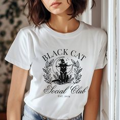 The "Black Cat Social Club Halloween T-Shirt" could feature a stylish and spooky design perfect for the Halloween season. Picture a sleek black cat with piercing eyes, sitting proudly atop a vintage, slightly distressed logo that reads "Black Cat Social Club" in a gothic font. The background could include subtle Halloween elements like crescent moons, stars, and perhaps a few bats flying around. The overall vibe would be mysterious and cool, perfect for anyone wanting to celebrate Halloween with Affordable Black Halloween T-shirt, Cat With Piercing, Be Mysterious, Bats Flying, Piercing Eyes, Halloween Elements, Gothic Font, The Black Cat, Spooky Designs