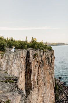 a man and woman standing on top of a cliff next to the ocean