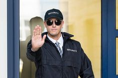 a security man in sunglasses and a black jacket is waving to the camera with his hand