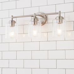 a bathroom light with three jars on the wall next to white brick walls and floor tiles