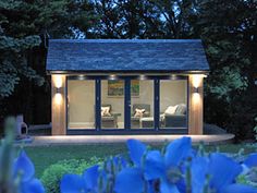 a small house with blue flowers in the foreground and trees in the background at night