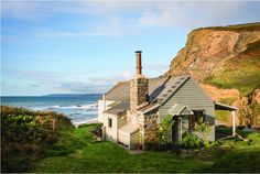 a small house sitting on top of a lush green hillside next to the ocean in front of a cliff