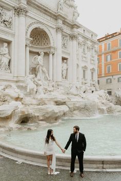 a man and woman holding hands in front of a fountain with statues on either side