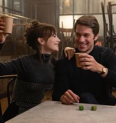 a man and woman sitting at a table with dice in front of them, drinking coffee