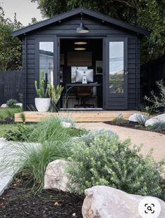 a small black shed sitting in the middle of a yard with rocks and plants around it