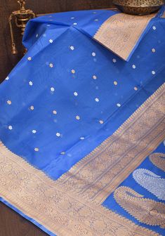 This Blue saree in pure Kora silk / Pure Organza from Banaras is adorned with gold and silver chunri butis and resplendent Elongated Paisley motifs that represent the fine craftsmanship and weaving heritage of Banaras! The Blue color serves as an amazing base for all the gold and silver zari work featured in the saree.  This drape could come to your rescue for a number of occasions from a simple family gathering to a big wedding. The fabric is sheer, mildly crisp and dreamy! SILK MARK CERTIFIED This saree is ready to wear with fall and pico done. Handmade silk tassels adorn the pallu and add more grace to it. An unstitched blouse fabric is included. *Note: There may be minor variations in the shade, the texture of the product. Hues/textures show differently due to variations in screen sett Traditional Blue Tissue Silk Sets, Blue Tissue Silk Blouse Piece For Transitional Season, Blue Tissue Silk Traditional Wear For Eid, Blue Silk Sets With Motifs, Blue Silk Traditional Wear With Motifs, Blue Elegant Paithani Silk Dupatta, Blue Tussar Silk Lehenga With Traditional Patterns, Blue Paithani Silk Saree Set, Blue Banarasi Silk Traditional Wear For Puja