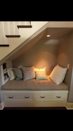 a room with some drawers under the stairs and pillows on top of it, in front of a stair case