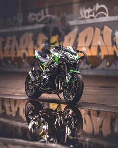 a green and black motorcycle parked in front of a graffiti covered wall with its reflection on the ground