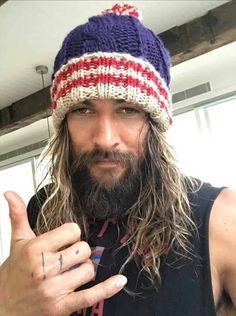 a man with long hair and a beard wearing a knitted hat gives the thumbs up sign