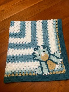 a blue and white crocheted blanket with a teddy bear on the bottom, sitting on a wooden floor