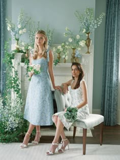 two women in dresses sitting next to each other on a chair with flowers and greenery behind them