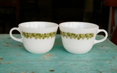 two white cups sitting on top of a wooden table next to each other with green leaves painted on them