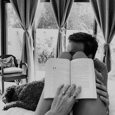 a woman reading a book while laying on top of a bed next to a window