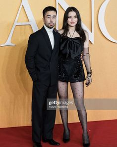 two people standing on a red carpet in front of an orange wall and one person wearing black