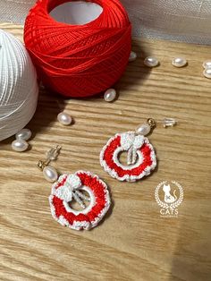 two red and white crochet earrings sitting next to balls of yarn on a table