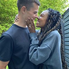 a man and woman standing next to each other in front of some trees with their faces close together