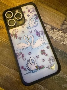 an iphone case with swans on it and two wedding rings in the middle, sitting on a wooden surface