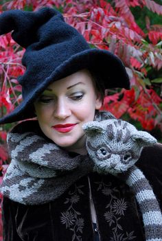 a woman wearing a black hat and scarf with a cat on her shoulder in front of red leaves