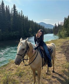 a woman riding on the back of a brown horse next to a river and forest