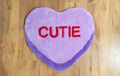 a heart shaped rug with the word cutie written on it, sitting on a wooden floor