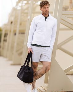 a man standing next to a wall holding a black handbag and wearing white shorts