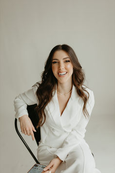 a woman sitting in a chair with her legs crossed and smiling at the camera she is wearing a white shirt and black pants